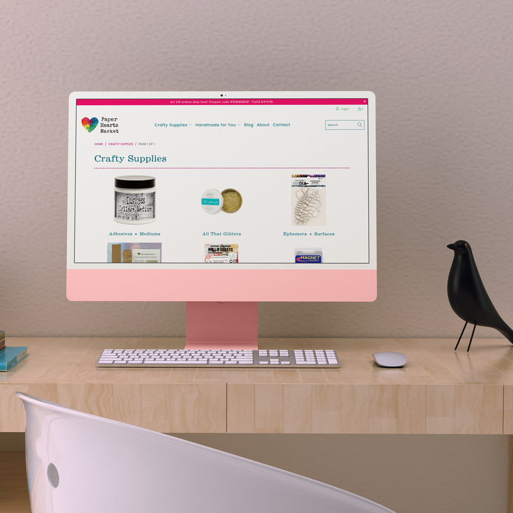 Mockup image of Paper Hearts Market Subcollection page on a pink desktop computer with a bird decor piece on a wooden desktop with a white retro chair in the foreground